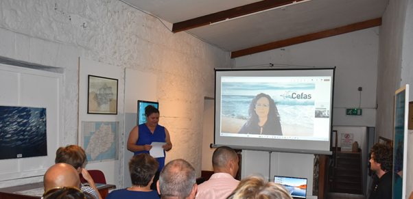 A women in a projection screen presents to a group of people sitting in a room. 