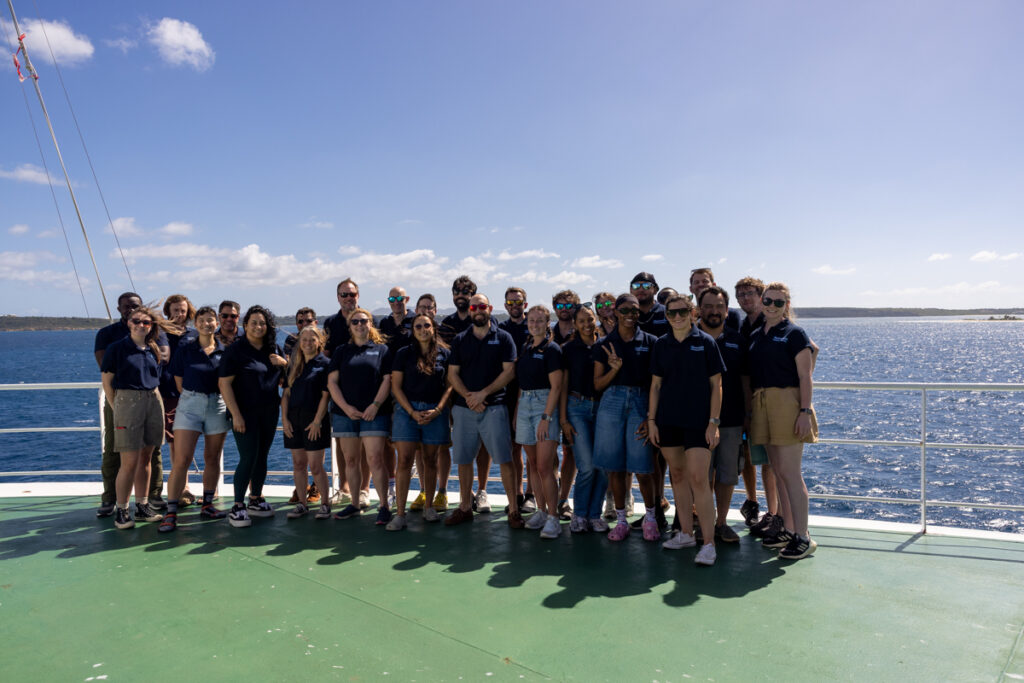 Group of people wearing navy polo shirts smiling on the bow of a ship with a green floor and blue, sunny skies.